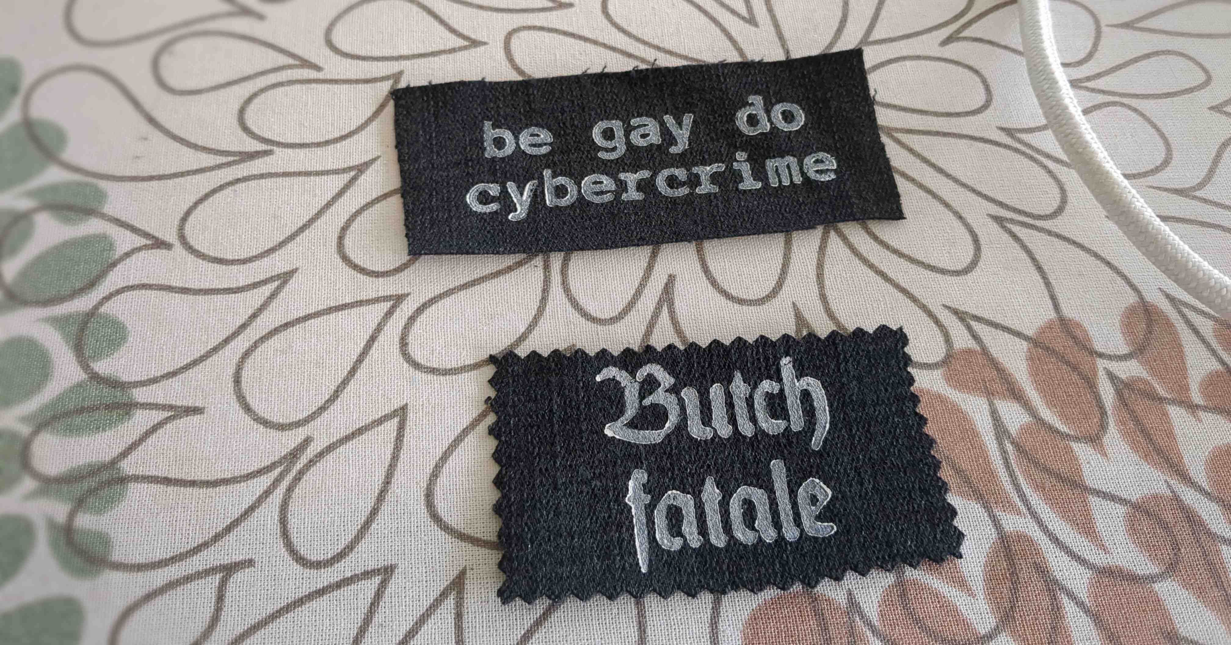 two patches on black denim. One says "be gay do cybercrime" and the other says "butch fatale"